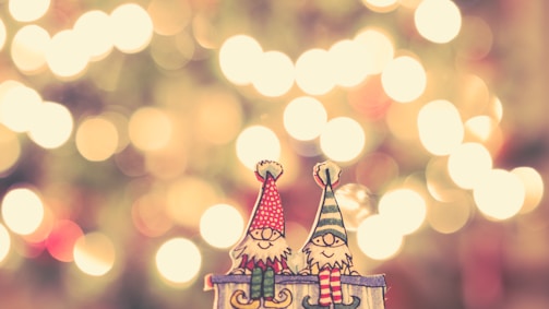 Two cartoon gnomes, one wearing a red hat and the other in a green and white striped hat, are sitting together on a ledge. They have cheerful expressions, surrounded by a backdrop of warm, soft light bokeh that creates a festive atmosphere.
