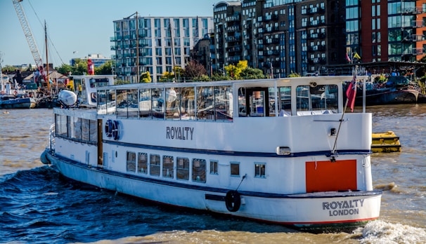 A passenger boat named 'Royalty London' is sailing on a river with modern buildings and a small harbor visible in the background. The boat is predominantly white and features clear windows with reflections. There are various structures on the shoreline, including apartment complexes and cranes, indicative of an urban riverside environment.