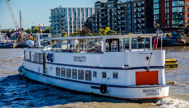 A passenger boat named 'Royalty London' is sailing on a river with modern buildings and a small harbor visible in the background. The boat is predominantly white and features clear windows with reflections. There are various structures on the shoreline, including apartment complexes and cranes, indicative of an urban riverside environment.