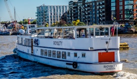 A passenger boat named 'Royalty London' is sailing on a river with modern buildings and a small harbor visible in the background. The boat is predominantly white and features clear windows with reflections. There are various structures on the shoreline, including apartment complexes and cranes, indicative of an urban riverside environment.