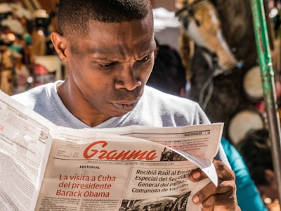 A man intently reads a newspaper titled 'Granma' outdoors, with articles about political events like a visit from Barack Obama. The background is slightly blurred, featuring various objects and people.