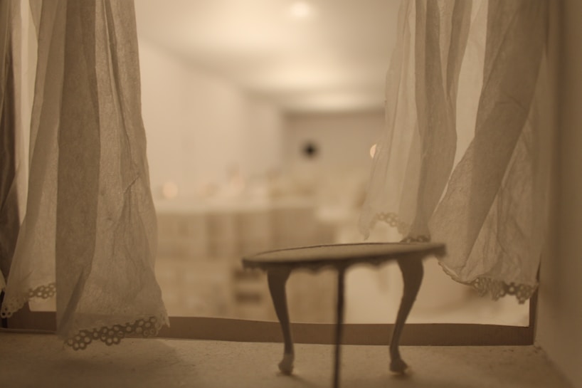 A softly lit room viewed through semi-transparent curtains, creating a gentle, dreamy ambiance. A small table with delicate legs is visible in the foreground, suggesting a miniature or abstract setting. The background is blurred, emphasizing a warm, inviting tone.