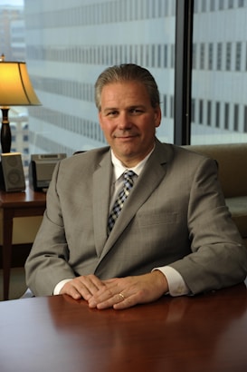 A man in a gray suit and checkered tie sits at a wooden desk in an office setting. The background features large windows with a view of tall office buildings. A table lamp and some office equipment are visible on a side table.