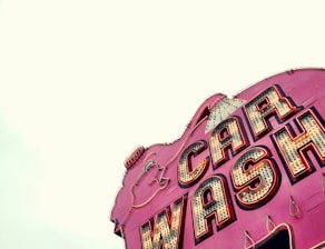 A vintage-style car wash sign with bold, illuminated letters. The sign has a retro aesthetic with large, outlined lettering on a vibrant pink background and incandescent bulbs.