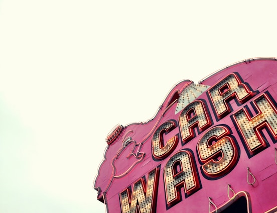 A vintage-style car wash sign with bold, illuminated letters. The sign has a retro aesthetic with large, outlined lettering on a vibrant pink background and incandescent bulbs.