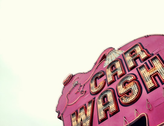 A vintage-style car wash sign with bold, illuminated letters. The sign has a retro aesthetic with large, outlined lettering on a vibrant pink background and incandescent bulbs.