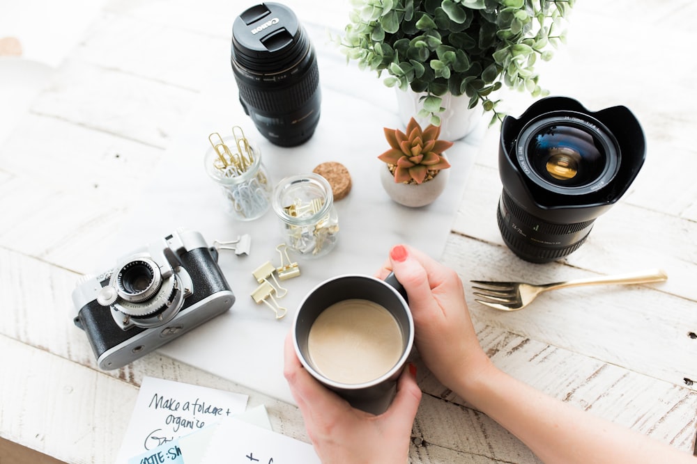 pessoa segurando caneca de café de cerâmica preta ao lado da câmera DSLR preta