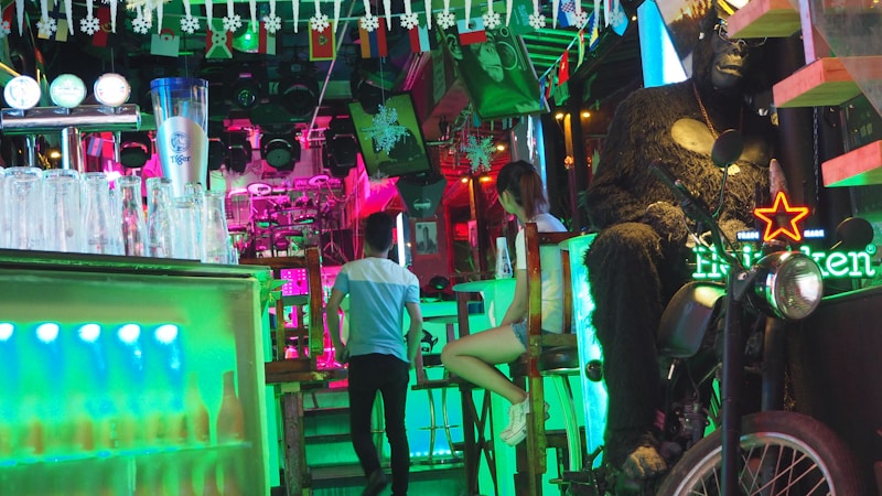 A vibrant and colorful bar setting with a mix of neon lights. Glasses and a large beer mug are on the counter to the left. In the background, a person in a light blue t-shirt is walking away and another person is sitting at the bar. On the right, a large gorilla statue sits on a motorcycle, with a Heineken neon sign nearby. The ceiling is decorated with small flags and hanging ornaments.