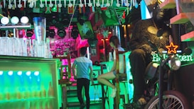 A vibrant and colorful bar setting with a mix of neon lights. Glasses and a large beer mug are on the counter to the left. In the background, a person in a light blue t-shirt is walking away and another person is sitting at the bar. On the right, a large gorilla statue sits on a motorcycle, with a Heineken neon sign nearby. The ceiling is decorated with small flags and hanging ornaments.