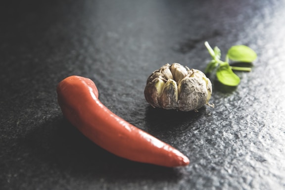 A red chili pepper lies on a dark textured surface next to a head of garlic. Nearby, a sprig of fresh basil with bright green leaves adds a touch of freshness.