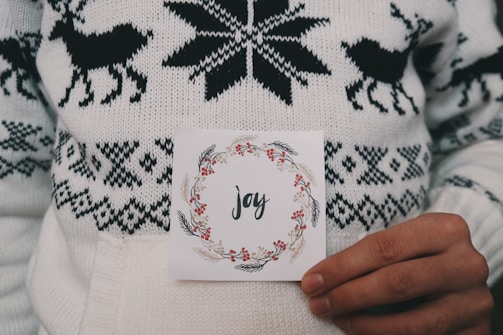 A person is holding a card with the word 'joy' written in elegant script, surrounded by a festive wreath design. The person is wearing a knitted white sweater featuring black reindeer and snowflake patterns.