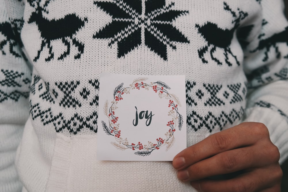 A person wearing a reindeer Christmas sweater while holding a card that says "Joy," with a wreath surrounding the writing.