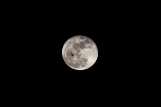 A bright, full moon captures attention in a dark night sky, with intricate details of its surface visible. A silhouette of an airplane is centrally positioned, creating a striking contrast against the illuminated lunar backdrop.