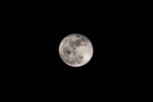 A bright, full moon captures attention in a dark night sky, with intricate details of its surface visible. A silhouette of an airplane is centrally positioned, creating a striking contrast against the illuminated lunar backdrop.