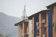 A series of brick buildings with blue accents and air conditioning units attached to the walls. An antenna tower is visible in the background, with a mountain range further behind. The sky is overcast, and some greenery is present at the bottom left corner.