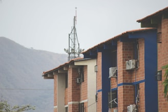 A series of brick buildings with blue accents and air conditioning units attached to the walls. An antenna tower is visible in the background, with a mountain range further behind. The sky is overcast, and some greenery is present at the bottom left corner.