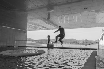 A person captured in mid-air, leaping over a shallow water feature with a patterned circular platform. The setting appears to be under an architectural structure with a reflective ceiling. In the background, a railing overlooks a body of water with a distant view of trees and a bridge.