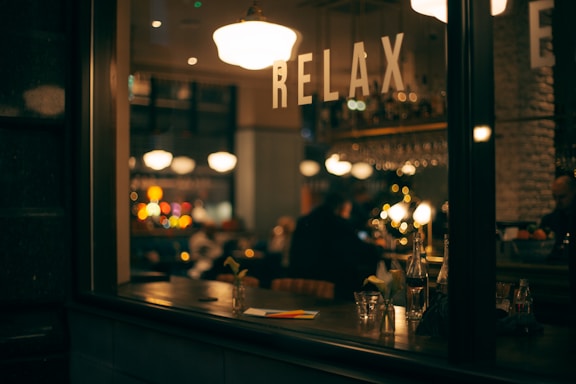 A cozy and warmly lit interior of a bar or café viewed through a window with the word 'RELAX' written on it. The atmosphere inside appears relaxed with patrons sitting and engaging in conversation. Bottles, glasses, and a small flower vase are visible on a counter, while soft, ambient lighting and blurred colorful lights contribute to a soothing ambiance.