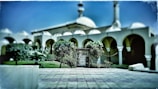 A mosque with distinctive arches and a dome, surrounded by greenery. The courtyard is paved and features a few trees and bushes, creating a symmetrical and serene setting.