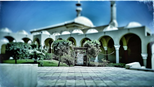 A mosque with distinctive arches and a dome, surrounded by greenery. The courtyard is paved and features a few trees and bushes, creating a symmetrical and serene setting.
