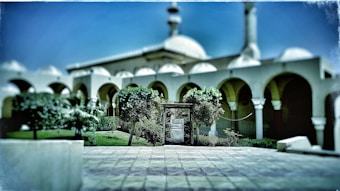 A mosque with distinctive arches and a dome, surrounded by greenery. The courtyard is paved and features a few trees and bushes, creating a symmetrical and serene setting.