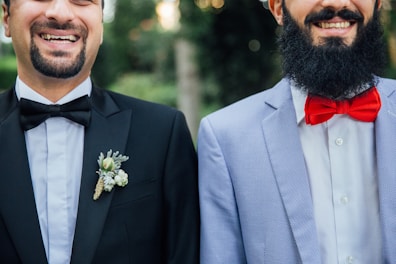 Two smiling men wearing formal attire, one in a black suit with a black bow tie and floral boutonniere, the other in a light blue suit jacket with a white shirt and bright red bow tie. Both have beards and are photographed from the chest up, standing side by side against a blurred green background.