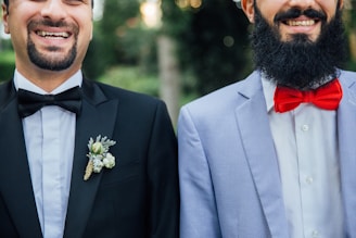 Two smiling men wearing formal attire, one in a black suit with a black bow tie and floral boutonniere, the other in a light blue suit jacket with a white shirt and bright red bow tie. Both have beards and are photographed from the chest up, standing side by side against a blurred green background.