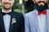 Two smiling men wearing formal attire, one in a black suit with a black bow tie and floral boutonniere, the other in a light blue suit jacket with a white shirt and bright red bow tie. Both have beards and are photographed from the chest up, standing side by side against a blurred green background.