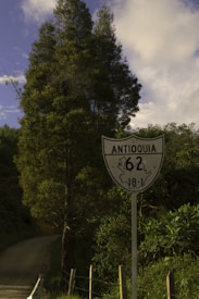 A road sign displays 'Antioquia 62 18-1' set against a backdrop of lush green trees and a dirt road leading into the distance. The sky is dotted with white clouds.
