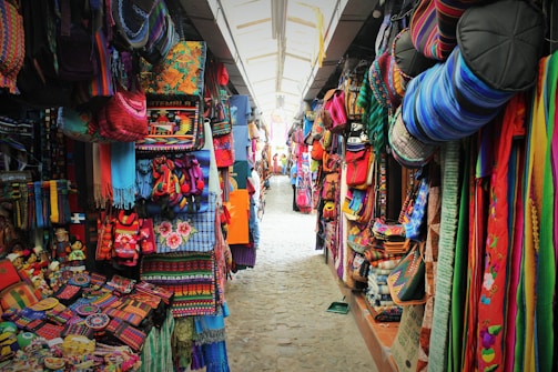 A vibrant market stall with colorful textiles, bags, and handcrafted items filling both sides of a narrow path. The ceiling is arched with a transparent covering, allowing light to illuminate the bright and diverse fabrics displayed in the market. Various patterns and designs are evident, showcasing a rich cultural aesthetic.