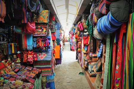 A vibrant market stall with colorful textiles, bags, and handcrafted items filling both sides of a narrow path. The ceiling is arched with a transparent covering, allowing light to illuminate the bright and diverse fabrics displayed in the market. Various patterns and designs are evident, showcasing a rich cultural aesthetic.