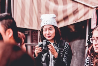 A person holding a camera is wearing a white beanie with spikes and a black leather jacket. They are surrounded by other people and standing in front of a striped canopy or tent.