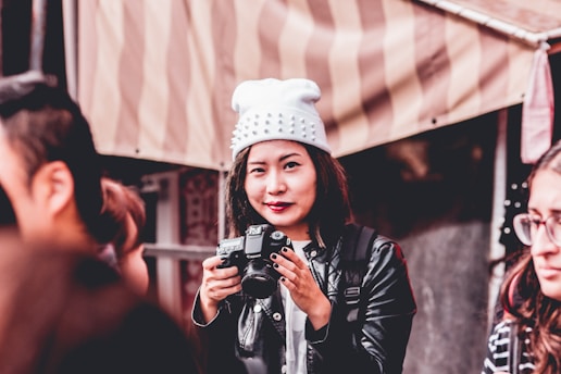 A person holding a camera is wearing a white beanie with spikes and a black leather jacket. They are surrounded by other people and standing in front of a striped canopy or tent.