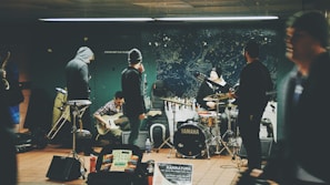 A group of musicians is performing in an indoor setting, likely a subway station. The ensemble includes a drummer, a guitarist, and a saxophonist. They are surrounded by various musical equipment, including drums marked with the Yamaha brand. The atmosphere is casual, with some members wearing beanies and hoodies. A mural with celestial motifs decorates the background wall.