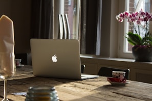 A sleek Apple MacBook open on a wooden desk with natural light streaming in.