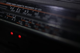 A close-up view of a vintage stereo radio cassette recorder. The focus is on the frequency display, which includes various numbers and units such as 540, 600, and 120m. Two small red indicator lights are glowing beneath the display, hinting at the device being powered on.
