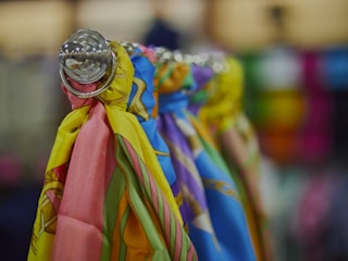 A close-up view of colorful scarves hanging on a rod, showcasing vibrant patterns and a glossy metal fixture. The background appears blurred, highlighting the focus on the fabric textures and hues.