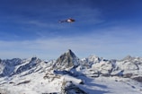 A red helicopter is flying over a range of snow-capped mountains under a clear blue sky. The landscape features rugged peaks and expansive areas of snow, with the helicopter carrying a long cable beneath it.