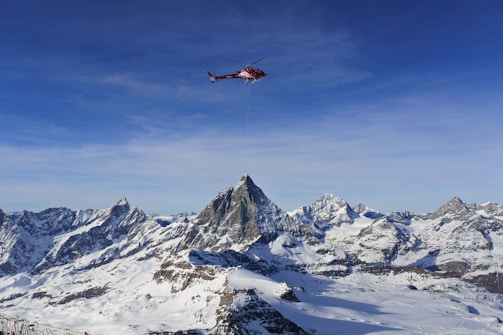 A red helicopter is flying over a range of snow-capped mountains under a clear blue sky. The landscape features rugged peaks and expansive areas of snow, with the helicopter carrying a long cable beneath it.