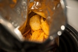 A partially opened bag of ridged potato chips reveals the crispy, golden snacks inside. The shiny silver interior of the bag reflects light, highlighting the appetizing texture and color of the chips. The view is slightly angled, focusing on the contents within.