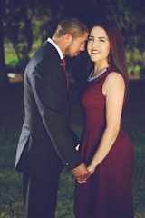 A man and a woman stand together in a grassy outdoor setting. The man, dressed in a dark suit and tie, leans his head affectionately towards the woman, who is wearing a maroon dress with a necklace. They are holding hands and appear close, surrounded by a blurred background of greenery.