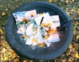 A black fire pit contains a pile of burning papers, including magazine pages and a 2016 calendar. The surrounding ground is covered in dry leaves and small patches of grass. Flames and partially burnt material are visible, along with orange-colored chips scattered among the papers.