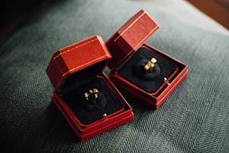 Two red velvet boxes are open to display elegant gold rings with intricate designs. The boxes are placed on a textured dark green fabric, creating a luxurious setting.