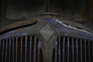 A close-up of an old, rusty car grille with the Renault logo prominently displayed in the center. The surface is covered in rust and peeling paint, giving it a weathered and aged appearance.