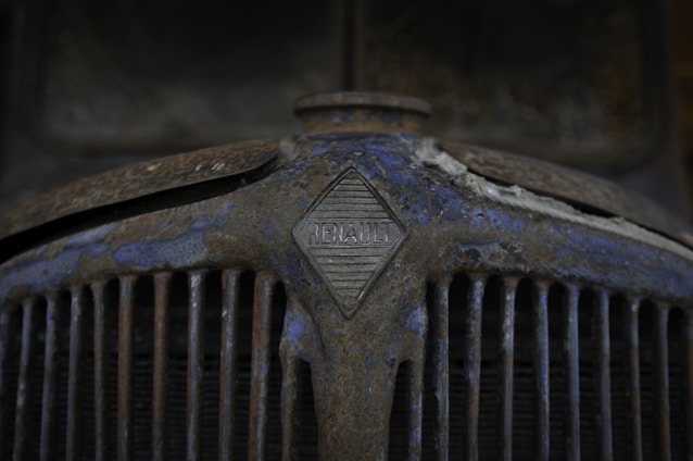 A close-up of an old, rusty car grille with the Renault logo prominently displayed in the center. The surface is covered in rust and peeling paint, giving it a weathered and aged appearance.