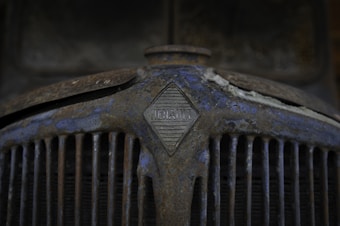 A close-up of an old, rusty car grille with the Renault logo prominently displayed in the center. The surface is covered in rust and peeling paint, giving it a weathered and aged appearance.
