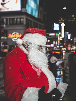 A person dressed as Santa Claus stands in a busy urban street at night. The scene is filled with bright city lights and illuminated signage in the background, with a bustling atmosphere.