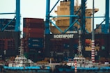 A large shipping port with numerous stacked cargo containers, cranes, and two small boats docked at the harbor. The containers are primarily red and blue and labeled with 'KMTC'. The cranes are towering, painted in blue, with the word 'NORTHPORT' prominently displayed. The scene is industrial, indicating a bustling area for shipping and logistics.