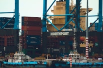 A large shipping port with numerous stacked cargo containers, cranes, and two small boats docked at the harbor. The containers are primarily red and blue and labeled with 'KMTC'. The cranes are towering, painted in blue, with the word 'NORTHPORT' prominently displayed. The scene is industrial, indicating a bustling area for shipping and logistics.
