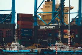 A large shipping port with numerous stacked cargo containers, cranes, and two small boats docked at the harbor. The containers are primarily red and blue and labeled with 'KMTC'. The cranes are towering, painted in blue, with the word 'NORTHPORT' prominently displayed. The scene is industrial, indicating a bustling area for shipping and logistics.